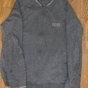 Grå ziptröja från Hugo Boss - Säljer en stilren grå ziptröja från Hugo Boss med diskret logga på bröstet. Tröjan har långa ärmar, ribbade muddar och dragkedja framtill. Perfekt för en avslappnad och snygg look.