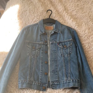 Klassisk jeansjacka från Levi's - Säljer en tidlös blå jeansjacka från Levi's med bruna knappar och två bröstfickor med lock. Jackan har klassisk krage och raka sömmar, perfekt till vår och sommar. Ikonisk röd Levi's-tagg på bröstfickan.