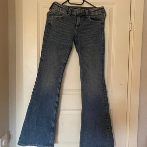 Blå bootcut jeans från H&M Divided - Säljer ett par klassiska blå bootcut jeans från H&M Divided. Jeansen har låg midja, fem fickor och snygga detaljer med knappar på bakfickorna. Perfekta för dig som gillar en retroinspirerad look med utsvängda ben.