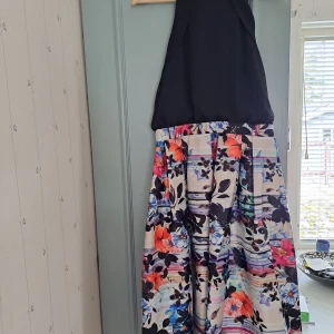 Svart och blommig långklänning med halterneck köpt i London - Säljer en långklänning med svart halterneck-topp och vid kjol med färgglatt blommigt mönster i orange, blått, rosa och lila. Kjolen har breda ränder och stora blommor, toppen knyts i nacken. Perfekt för sommarens tillfällen! Från spotlight i London 