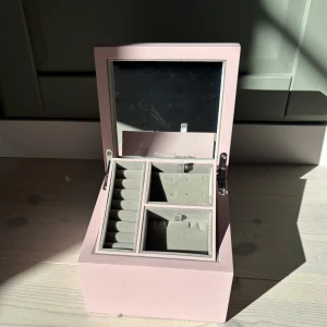 Rosa smyckeskrin från Pandora - Säljer ett stilrent rosa smyckeskrin från Pandora med spegel i locket och flera fack för ringar, örhängen och andra smycken. Perfekt för att organisera dina favoritsmycken på ett snyggt sätt. Skrinet har två våningar och en mjuk insida i grå sammet.