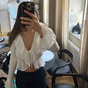 Vit omlottblus med volang - Säljer en gullig vit omlottblus med volangdetaljer och knyt framtill. Blusen har långa ärmar i ett lätt, transparent material och en djup v-ringning.💓