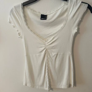 Vit topp med spetsdetaljer  - Söt vit topp från Gina tricot med v-ringning, spetskant och rynk framtill. Korta ärmar och mjukt, stretchigt material. Perfekt för en fräsch och feminin look.
