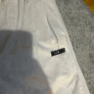 Beige mjukisbyxor från Moschino - Säljer ett par beige mjukisbyxor från Moschino med broderad logga på ena benet. Byxorna har resår i midjan och är tillverkade i ett mjukt material som känns riktigt skönt mot huden. Perfekta för en avslappnad stil.