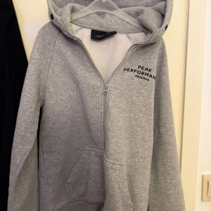 Grå hoodie från Peak Performance - Säljer en grå hoodie från Peak Performance med dragkedja och huva. Tröjan har fickor framtill och logga på bröstet. Perfekt för en avslappnad stil och enkel att matcha med det mesta.
