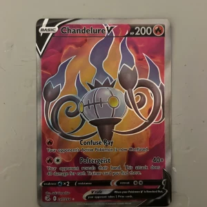 Chandelure V Pokémonkort - Säljer ett Chandelure V Pokémonkort med 200 HP. Kortet har en färgstark design i rött, lila och gult med holografisk effekt. Det är ett basic fire-type kort med attacker som Confuse Ray och Poltergeist. Perfekt för samlare eller Pokémon TCG-spelare.