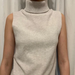 Beige ärmlös stickad polotröja - Säljer en beige ärmlös stickad polotröja med hög krage. Perfekt att styla lager på lager eller bära som den är. Mjuk och skön med ribbad polokrage för extra stilpoäng.