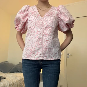 Vit blus med rosa rosor och puffärmar - Säljer en söt vit blus med rosa rosmönster och stora puffärmar. Blusen har v-ringning och korta ärmar som ger en romantisk känsla. Perfekt att styla med jeans eller kjol för en trendig look.