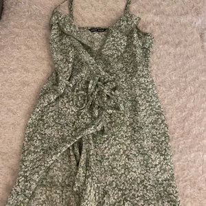 Grön blommig omlottklänning med volang - Säljer en söt tunn grön omlottklänning med vitt blommönster. Klänningen har smala axelband, volangdetaljer längs urringningen och knyt i midjan. 