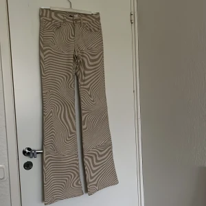 Beige mönstrade byxor från Divided - Säljer ett par beige byxor från Divided med unikt, vågigt mönster. Byxorna har bootcut-modell och normal passform. aldrig använda.🤍🤍storlek 32