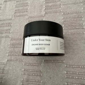 Oöppnad Under Your Skin Organic Body Scrub - En burk med Under Your Skin Organic Body Scrub, 200 ml. Kommer i en svart rund plastburk med vit etikett. Passar alla hudtyper och är ekologisk. Perfekt för att exfoliera huden och ge en fräsch känsla.
