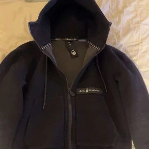 Svart hoodie från Sail Racing - Säljer en svart hoodie från Sail Racing med dragkedja och huva. Jackan har två stora fickor framtill och justerbar dragsko i huvan. Perfekt för svala dagar och snygg logga på bröstet.