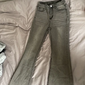 Grå flare jeans med resårmidja - Säljer ett par gråa flared jeans med resår i midjan och klassiska fickor fram och bak. Jeansen har en knapp och dragkedja framtill och är i stretchigt material. Perfekta för dig som gillar en avslappnad men trendig look. 