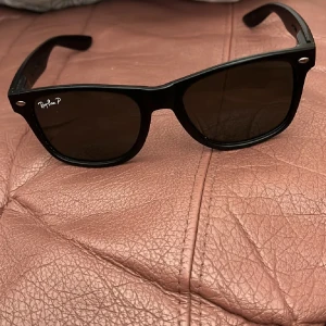 Svarta solglasögon från Ray-Ban - Klassiska svarta solglasögon från Ray-Ban med Wayfarer-inspirerad form. Glasögonen har mörka linser och diskret logga på ena linsen. Passar perfekt för en stilren look.