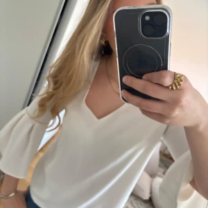 Vit blus med volangärmar - Säljer en stilren vit blus med v-ringning och vida volangärmar. Perfekt att styla med jeans eller kjol för en trendig look. Blusen har en lös passform och är tillverkad i ett mjukt material. Storlek S