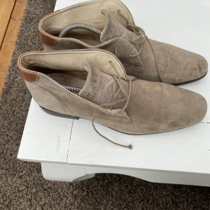 Beige mockaboots från Marville - Säljer ett par beige boots i mocka från Marville med snörning och rund tå. Skorna har en liten brun detalj vid hälen och platt sula. Perfekta för dig som gillar stilrena och klassiska skor.