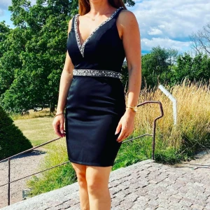 Svart kortklänning med glittriga detaljer - Säljer en svart kortklänning med v-ringning och glittriga detaljer längs urringning och midja. Klänningen har breda axelband och en figurnära passform som framhäver siluetten. Perfekt för festliga tillfällen!