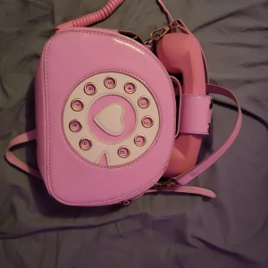 Rosa axelväska med telefon-design - Superunik rosa axelväska formad som en gammal telefon med snurrskiva och hjärtdetalj. Väskan har axelrem och dragkedja, perfekt för dig som vill sticka ut med en lekfull accessoar. Materialet är blankt och känns som syntet.