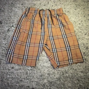 Beige rutiga shorts från Burberry - Säljer ett par klassiska beige shorts från Burberry med det ikoniska rutiga mönstret i svart, vitt och rött. Shortsen har resår i midjan och snörning för extra komfort. Perfekta för sommaren och har en avslappnad passform.