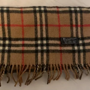 Beige rutig halsduk från Burberry - Klassisk beige halsduk från Burberry med rutigt mönster i svart, vitt och rött. Gjord i 100% lammull och har fransar i kanterna. Perfekt accessoar för att lyfta din outfit.
