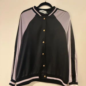 Svart och lila bomberjacka från Moves by Minimum - Säljer en svart och lila bomberjacka från Moves by Minimum. Jackan har guldiga knappar framtill, randiga muddar och sportig design. Tillverkad i polyester med elastan för extra komfort. Perfekt för dig som gillar en avslappnad stil.