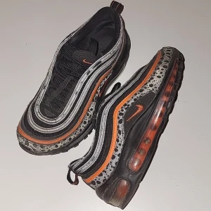 Nike Air Max 97 med orange detaljer - Säljer ett par Nike Air Max 97 med svart bas, orange swoosh och detaljer samt reflekterande silverränder. Skorna har synlig luftbubbla i sulan och ett unikt mönster runt sulkanten. Perfekta för dig som vill sticka ut med dina sneakers. Användningen syns mest på skosnören.