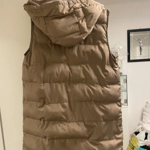 Beige dunväst med huva - Säljer en beige dunväst med huva och dragkedja framtill. Västen har två fickor med dragkedja och är vadderad för extra värme. Perfekt för lager-på-lager under kyliga dagar. Oanvänd 