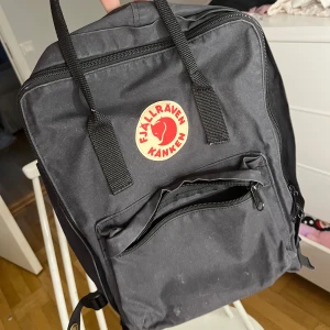 Svart ryggsäck från Fjällräven Kånken - Klassisk svart Fjällräven Kånken ryggsäck med ikonisk röd logga framtill. Väskan har ett stort huvudfack, en mindre ficka med dragkedja på framsidan och två sidofickor. Praktiska handtag upptill och justerbara axelremmar. Säljer billigare d det är defekter på den! Ny pris 1200 säljer för 400