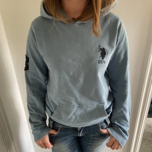 Ljusblå hoodie från USPA - Säljer en ljusblå hoodie från USPA med broderad logga på bröstet  och på högra axeln och en ficka framtill. Tröjan har huva och långa ärmar, perfekt för en avslappnad stil. Passar dig som gillar klassiska och bekväma plagg. Storleken 14-15 men passar fint pp mig som normalt har size small