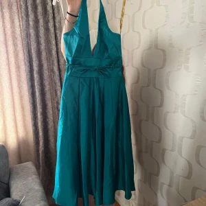 Turkos halterneckklänning med rosettdetalj - Säljer en turkos halterneckklänning med djup v-ringning och rosettdetalj i midjan. Klänningen har ett elegant fall och är tillverkad i ett glansigt material som ger en lyxig känsla. Perfekt för bal eller festliga tillfällen.