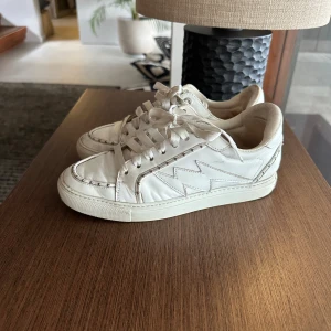 Vita sneakers från Zadic & Voltaire - Säljer ett par vita sneakers från zadic & voltaire med coola silverfärgade nitar längs sidorna och en blixtdetalj. Skorna har snörning och är tillverkade i skinn med rund tå och platt sula. Perfekta för dig som vill ha en stilren men edgy look.