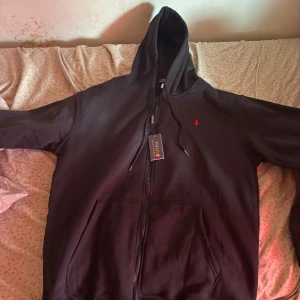 Svart hoodie från Polo Ralph Lauren - Säljer en svart hoodie från Polo Ralph Lauren med dragkedja och huva. Klassisk röd logga broderad på bröstet och två fickor framtill. Perfekt för en avslappnad stil och enkel att matcha med det mesta. Det står 3XL fast passar nog nån med storlek XXL. Pris kan diskuteras