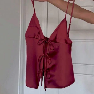 Vinröd satinlinnetopp  - Säljer en vinröd linnetopp i satin med smala axelband och tre dekorativa knyt framtill. Toppen har en djup v-ringning och justerbara axelband för bästa passform. Jätte snygg men har inte kommit till användning💕