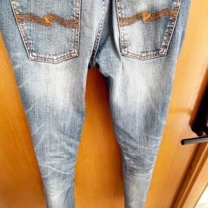 Blå jeans från Nudie Jeans - Säljer ett par klassiska blå jeans från Nudie Jeans med snygga slitningar och kontrastsömmar. Bakfickorna har Nudie Jeans ikoniska broderade vågmönster. Perfekta för dig som gillar en tidlös denimlook.                                       Nypris ~1600kr                                                                           Hör av dig! Alla frågor är välkomna! Går gärna ner i pris vid köp av bundles!