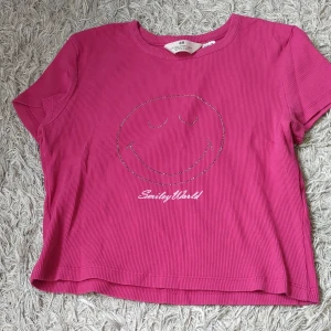 Rosa croppad t-shirt med smiley - En rosa croppad t-shirt från HM med ett glittrigt smiley-ansikte och texten 'SmileyWorld' framtill. T-shirten har korta ärmar och rund hals. Perfekt för en färgglad vardagslook. Skön att ha hemma bara.