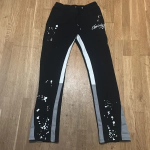 Svarta mjukisbyxor med vita färgstänk och gråa paneler - Säljer ett par svarta mjukisbyxor med gråa paneler längs benen och vita färgstänk som detalj. Byxorna har snörning i midjan och tryckt text på ena benet. Perfekta för en avslappnad stil.