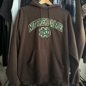 Brun Notre Dame hoodie - Säljer en brun hoodie med grön och vit Notre Dame-logga på bröstet. Tröjan har huva med dragsko och en stor magficka. Perfekt för dig som gillar college-stil och vill ha något bekvämt och snyggt.
