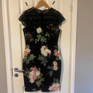 Svart blommig spetsklänning - Säljer en svart klänning med blommigt mönster och fina spetsdetaljer upptill. Klänningen har korta ärmar och en figurnära passform som framhäver siluetten. Perfekt för dig som vill ha en elegant och romantisk look. Använd 1 gång. Paperdoll klänning☺️