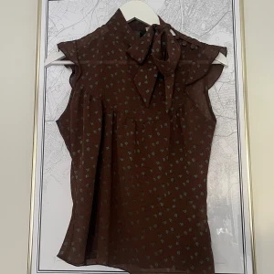 Brun blus med rosett och småblommigt mönster - Vintage blus i mesh med små blå blommor och knytrosett vid halsen (kan knytas upp). Blusen är ärmlös och har dekorativa knappar på axeln. Perfekt för sommaren 🌹