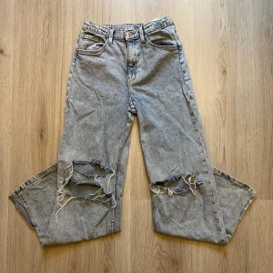 Blå jeans med slitningar - Säljer ett par ljus blåa vida jeans från H&M med hög midja och slitningar över båda knäna. Jeansen har klassisk femficksdesign. Dom ändå defekterna som finns är trådarna vid knäna som gått av. Skriv om du har frågor o läs helst min bio innan du köper🫶🏻🫶🏻