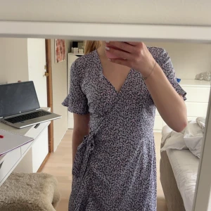 Ljuslila blommig omlottklänning - Säljer en söt ljuslila omlottklänning med småblommigt mönster. Klänningen har korta volangärmar, knyt i midjan och v-ringad hals. Perfekt för vår och sommar! Aldrig kommit till användning! Pris går att diskutera