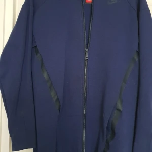 Blå zip-hoodie från Nike - Mörkblå hoodie från Nike med hel dragkedja framtill och svarta detaljer längs sidorna. Jackan har huva och långa ärmar, perfekt för träning eller chill. Klassisk Nike-logga på bröstet och sportig look. Stor i stolek passar inte mig som har xs-s