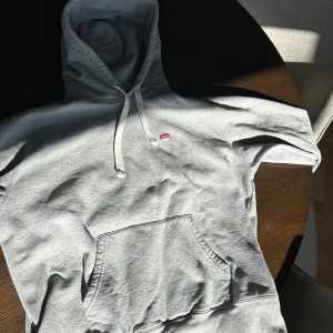 Grå hoodie från Supreme - Säljer en klassisk grå hoodie från Supreme med liten röd logotyp på bröstet. Tröjan har huva med snören och en stor magficka framtill. Perfekt för en avslappnad stil. Storlek Medium men passar även S. Äkthetsbevis finns.