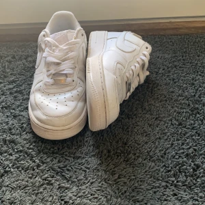 Vita Nike Air Force 1 sneakers - Säljer ett par klassiska vita Nike Air Force 1 sneakers med låg profil. Skorna har rund tå, snörning och är tillverkade i skinn med vit sula och diskret Nike-logga på sidan. Perfekta för en clean och stilren look.