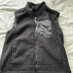 Svart fleeceväst  - Säljer en svart fleeceväst med hög krage och hel dragkedja framtill. Västen har en praktisk bröstficka i syntetmaterial med dragkedja. Perfekt för lager-på-lager under kyliga dagar. Priset är inte hugget i sten!