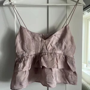 Ljusrosa volangtop med tunna axelband - Säljer en ljusrosa croppad topp med tunna axelband och dubbla volanger. Toppen har knappar framtill och är tillverkad i ett glansigt material som ger en lyxig känsla. Perfekt för sommardagar eller festliga tillfällen. Skriv för fler bilder💞