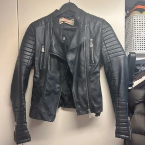 Svart skinnjacka  - Säljer en svart skinnjacka i storlek 38. Jackan har dragkedjor framtill, på ärmarna och vid fickorna samt quiltade detaljer på axlar och ärmar. Snygg bikerstil med markerade sömmar och snörning bak.