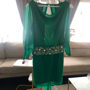 Grön klänning med pärldetaljer från S.Y.L - Säljer en smaragdgrön klänning från S.Y.L med transparenta långa ärmar och bred midja dekorerad med pärlor och paljetter. Klänningen har en elegant, lös passform och dragkedja bak. Perfekt för speciella tillfällen!