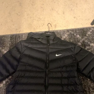 Svart dunjacka från Nike - Säljer en svart dunjacka från Nike med huva och dragkedja framtill. Jackan har en klassisk pufferdesign och Nike-logga på bröstet. Perfekt för kyligare dagar.