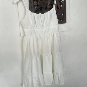 Vit somrig klänning från SHEIN - Söt vit klänning från SHEIN med smala axelband och volangdetaljer. Klänningen har en lätt rynkad struktur och är sydd i flera lager för en luftig känsla. Perfekt för varma dagar och har en feminin, flowy siluett.